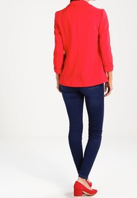 Blazer rouge structuré avec des manches retroussées, associé à un jean bleu foncé. Le mannequin porte des talons rouges, mettant en valeur une silhouette ajustée.