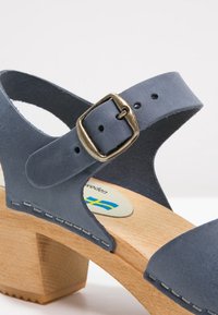 Sandale en daim bleu avec boucle en laiton réglable, talon bloc en bois, et étiquette du drapeau suédois sur la semelle intérieure.