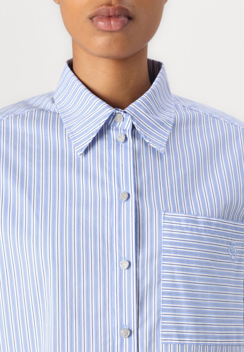 Koszula w paski button-up w jasnoniebieskim i białym kolorze. Posiada zaostrzony kołnierzyk, srebrne guziki oraz kieszeń na piersi z haftowanym logo.