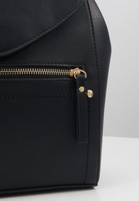 Černý batoh z umělé kůže s kulatou klopou, zlatým zipem, boční kapsou a vystupujícími švy. Hladký povrch a minimalistický design.