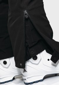 Pantalon noir avec une fermeture éclair latérale partiellement ouverte sur des bottes blanches montantes avec des détails circulaires noirs sur les côtés.
