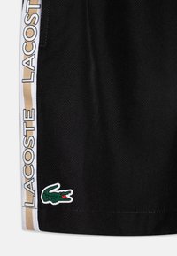 Shorts Lacoste noirs avec une surface texturée, rayures latérales blanches et dorées, et un patch de crocodile vert près de l'ourlet.
