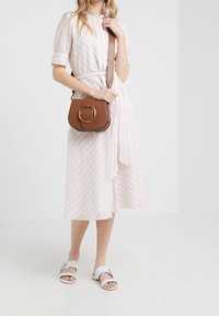 Ljusrosa och vit randig klänning med bälte vid midjan, tillsammans med en brun handväska med en gyllene cirkulär detalj och vita flata sandaler.