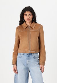 Brun semsketjakke med tilbehør av nagler, klassisk krage og glidelås foran. Båret med avslappede lys blå jeans.