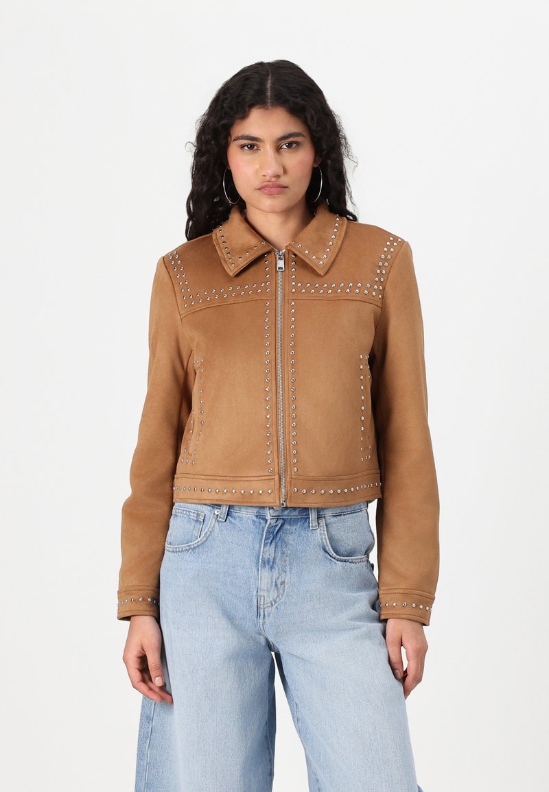 Brun semsketjakke med tilbehør av nagler, klassisk krage og glidelås foran. Båret med avslappede lys blå jeans.