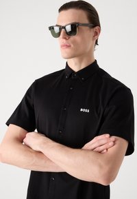 Juodas trumpų rankovių mygtukinis marškinėliai su apykakle; turi baltą "BOSS" logotipą; modelis dėvi tamsius akinius nuo saulės su žaliais objektyvais ir juodais rėmeliais.