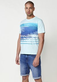 Ljusblå bomulls t-shirt med en tropisk design featuring palmblad i olika blå nyanser, kombinerad med uppvikta blå denimshorts.