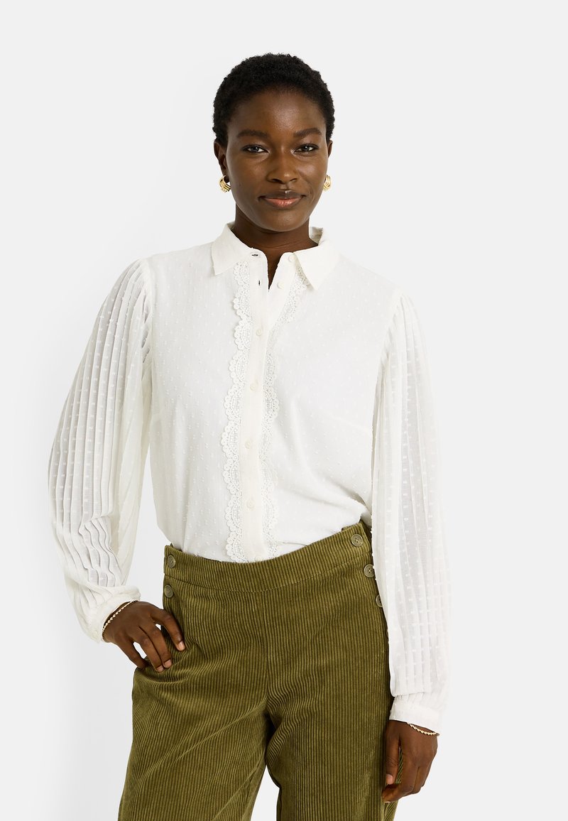 Blusa blanca con rayas texturizadas, borde de encaje en la parte delantera y puños abotonados. Combinada con pantalones de pana color verde oliva con botones laterales.