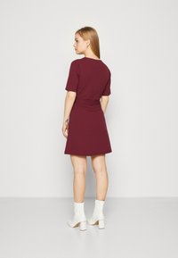 Burgundy színű ruha rövid ujjal, szűkített derékkal és bővülő szoknyával. A modellen fehér magassarkú bokacsizma van; sima anyagú kivitel.