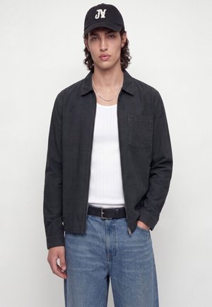 Jeune homme portant une casquette noire, une veste zippée foncée sur un débardeur blanc côtelé, un jean bleu clair et une ceinture noire, debout sur un fond blanc.