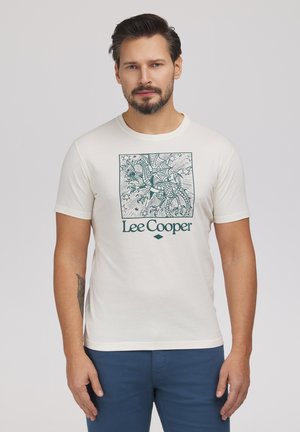 Mężczyzna z brodą w białej koszulce Lee Cooper z szczegółową grafiką smoka, w niebieskich spodniach, stojący na jednolitym tle.