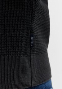 Pull noir texturé avec un motif tissé et un ourlet côtelé. Présente une petite étiquette bleue "MUSTANG" sur le côté.