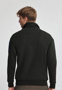 Svart sweatshirt med hög krage, ribbade ärmslut och sidodetaljer i mörkblått. Tyget verkar mjukt med en enkel design.