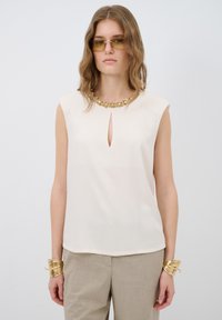 Blusa sin mangas color crema con escote en forma de llave y cuello adornado. Tejido suave con un ajuste relajado; los acentos dorados complementan el diseño.