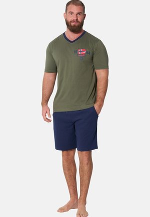 Bärtiger Mann steht barfuß, trägt ein olivgrünes T-Shirt mit einer rot-weißen Flaggen-Grafik und dunkelblaue Shorts, die Hände an den Seiten.