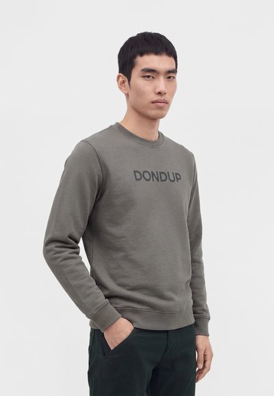 Млад мъж, облечен в сив суичър с текст "DONDUP" и тъмни панталони, стои с ръка в джоба си пред едноцветен фон.