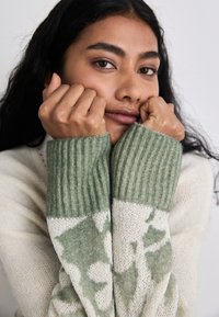 Strickpullover in Creme mit grünem Blattmuster, ausgestattet mit gerippten grünen Bündchen. Die Textur wirkt weich und gemütlich mit einer lockeren Passform.