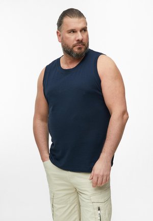 Man met baard en samengebonden haar, met een marineblauw mouwloos shirt en beige cargoshorts, staand met handen in de zakken, kijkend naar de zijkant.