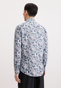 Camicia bianca a maniche lunghe con un motivo floreale nelle tonalità di blu, verde e rosso. Presenta un colletto con bottoni e un orlo curvo. Tessuto dalla consistenza liscia.