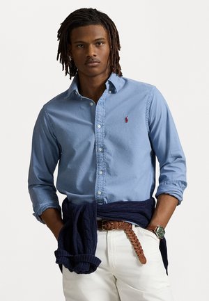 Lichtblauw button-down overhemd met een klein rood logo, opgerolde mouwen, gecombineerd met witte broek en een marineblauwe trui om de taille geknoopt. Bruin gevlochten riem.
