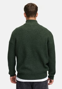 Mann iført en mørkegrønn ribbestrikket genser med høy krage, st�år med ryggen til kameraet, over en hvit skjorte og mørke jeans.