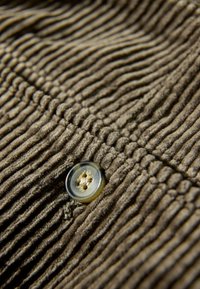 Bruine corduroy stof met smalle, verticale strepen en een grote, ronde, lichtgekleurde knop met vier gaten. Gelaagdheidsdetail zichtbaar.