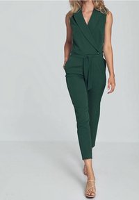 Grüner ärmelloser Jumpsuit mit tiefem Ausschnitt, Gürtel in der Taille, seitlichen Taschen, schmal zulaufenden Beinen und glatter Stoffbeschaffenheit.