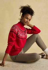 Red knit sweater with snowflake pattern on yoke, high neck, and ribbed cuffs, paired with checkered pants and light-colored shoes.