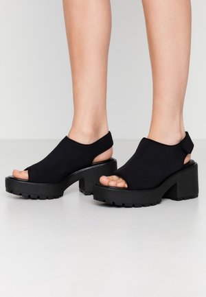 Mustat avokkaat, joissa on paksut lohko- ja takaremmit, käytössä paljain jaloin neutraalia taustaa vasten.