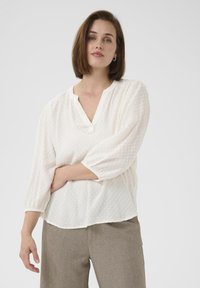 Blusa blanca texturizada con escote en V, mangas abullonadas de tres cuartos y dobladillo ligeramente curvado. Combinada con pantalones ajustados de color marrón claro.