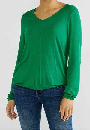 Kobieta ubrana w zieloną bluzkę z długim rękawem i lekko wyciętym dekoltem w serek oraz niebieskie dżinsy, stojąca z rozluźnionymi rękami na neutralnym tle.