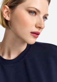 Maglietta blu navy a girocollo con una superficie liscia; primo piano di un volto con capelli biondi corti e un trucco sobrio con labbra rosa.