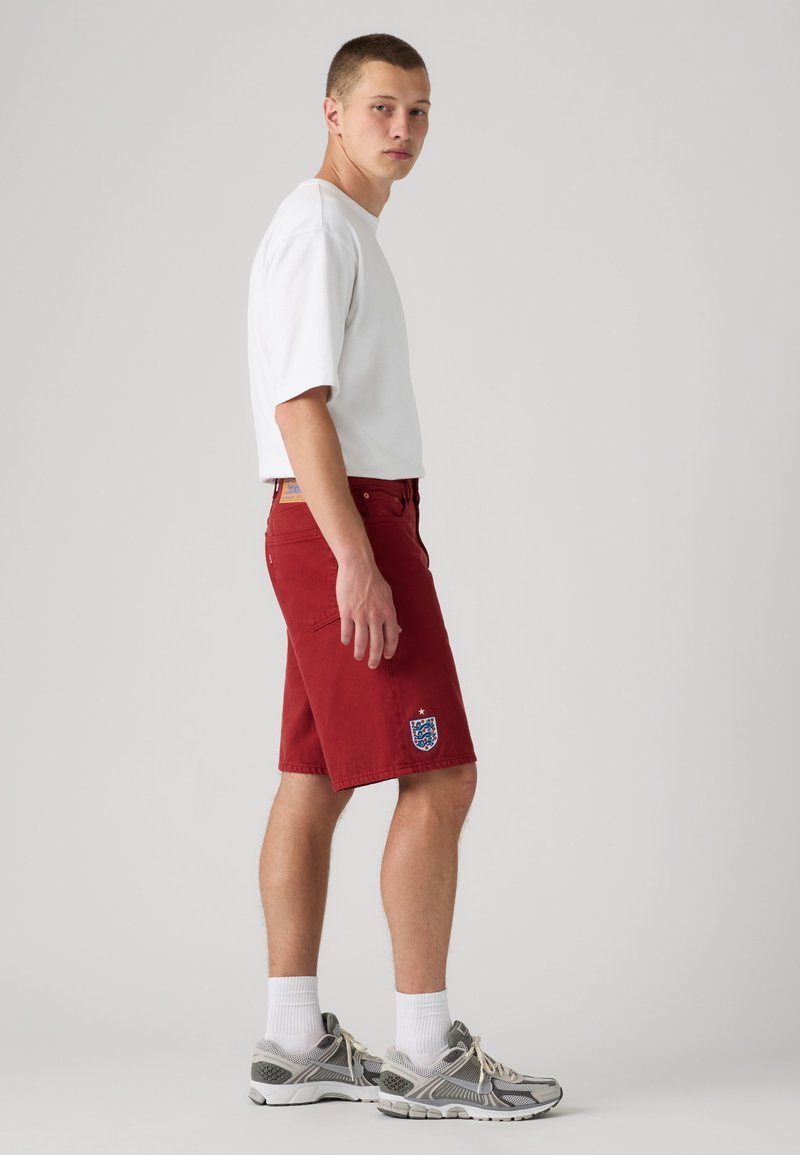 Joven de pie de lado vestido con una camiseta blanca, pantalones cortos rojos con el escudo de fútbol de Inglaterra, calcetines blancos y zapatillas grises.