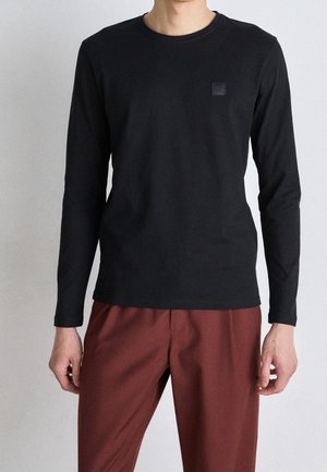 Personne portant un t-shirt noir à manches longues et un pantalon bordeaux, debout devant un fond uni clair.