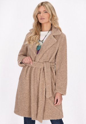 Donna con lunghi capelli biondi che indossa un cappotto teddy marrone chiaro con cintura sopra una maglietta bianca e una collana di perline su uno sfondo bianco semplice.