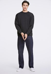 Sudadera negra de cuello redondo con hombros caídos, combinada con pantalones de mezclilla azul oscuro. El modelo lleva zapatillas blancas, con un fondo neutro.