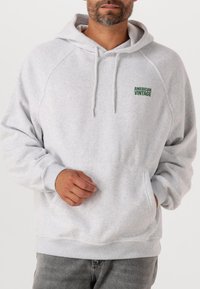 Hoodie gris clair en matière douce, doté d'une poche kangourou, d'une capuche avec cordon de serrage et du logo vert "American Vintage" sur la poitrine.