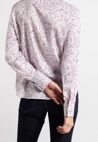 Persona che indossa una blusa bianca a maniche lunghe con punti rossi e neri, le mani dietro la schiena, abbinata a jeans blu scuro, in piedi contro uno sfondo neutro.