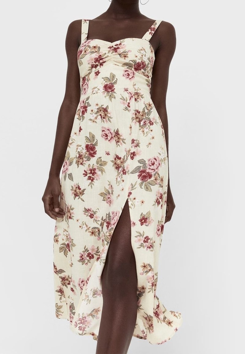 Robe midi à fleurs avec une base crème, arborant des motifs floraux roses et verts, fente latérale et fines bretelles. Tissu doux et léger.