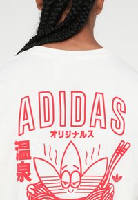 Balta T-krekls ar lielu sarkanu grafiku, kurā ir "ADIDAS" un karikatūras dizains ar bļodu nūdeles, papildināts ar kociņiem un Adidas logo.