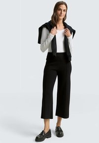 Zwarte cropped broek met een hoge taille, gecombineerd met een wit langemouwig shirt. Een zwarte trui hangt over de schouders. Zwarte loafers maken de look compleet.