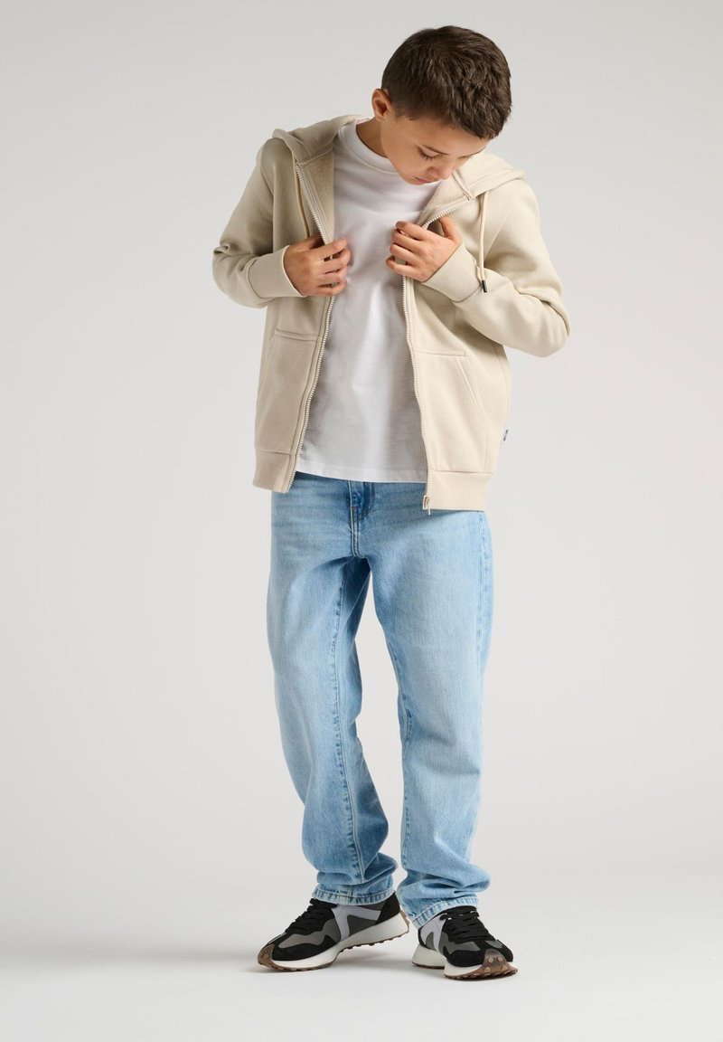 Un garçon portant une veste zippée beige, un t-shirt blanc, un jean bleu clair et des baskets noir et blanc, regardant vers le bas tout en ajustant sa veste.