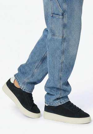 Persona che indossa jeans azzurri chiari e scarpe da ginnastica nere con suola bianca, in piedi su uno sfondo bianco.