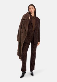 Brown patterned suit with a herringbone design, paired with a brown shirt and oversized, textured brown coat. Black heeled ankle boots.