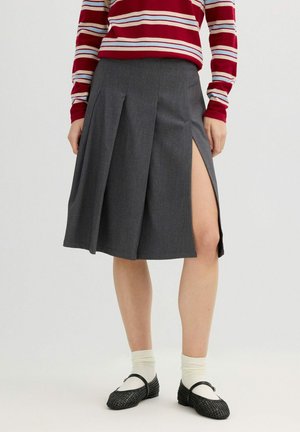 Mujer con falda gris hasta la rodilla con una abertura lateral, camiseta de rayas rojas y blancas, calcetines blancos y zapatos negros con tiras entrelazadas.