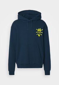 Tummansininen huppari, joka on valmistettu pehmeästä kankaasta. Siinä on kengurupocket ja keltainen graafinen kuva hymyilevästä ranskanperunamaljasta vasemmassa rinnassa.