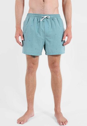 Hombre con pantalones cortos de baño azul claro con pequeños patrones abstractos y un cordón blanco, de pie descalzo frente a un fondo liso.