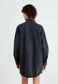 Donkergrijze denim shirtjurk met een kraag, lange mouwen, knoopmanchetten en een gebogen zoom. Licht oversized pasvorm met minimale stikdetails.