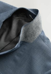 Niebieska teksturowana kurtka z szarym filcowym kołnierzem. Wnętrze posiada ciemny wzorzysty podszewki. Skupia się na nowoczesnym designie i kontraście materiałów.