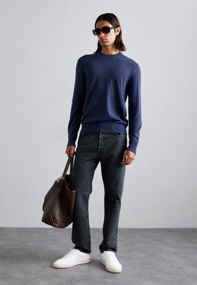 Person wearing navy sweater, black jeans, white sneakers, black sunglasses, holding large brown leather bag, standing on gray floor against white wall.